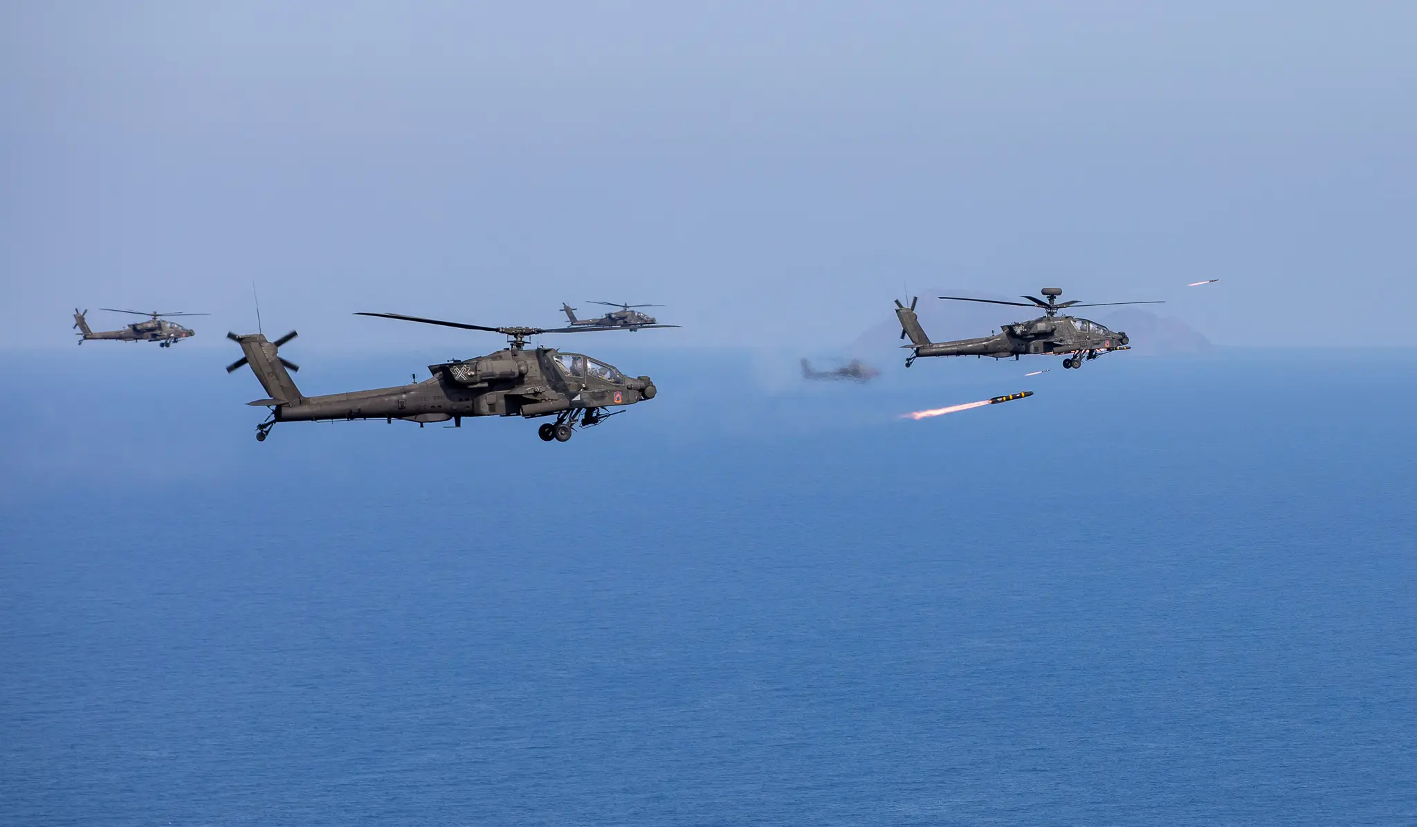 AH-64D Apache Longbows in formation over the sea as one launches a Hellfire missile